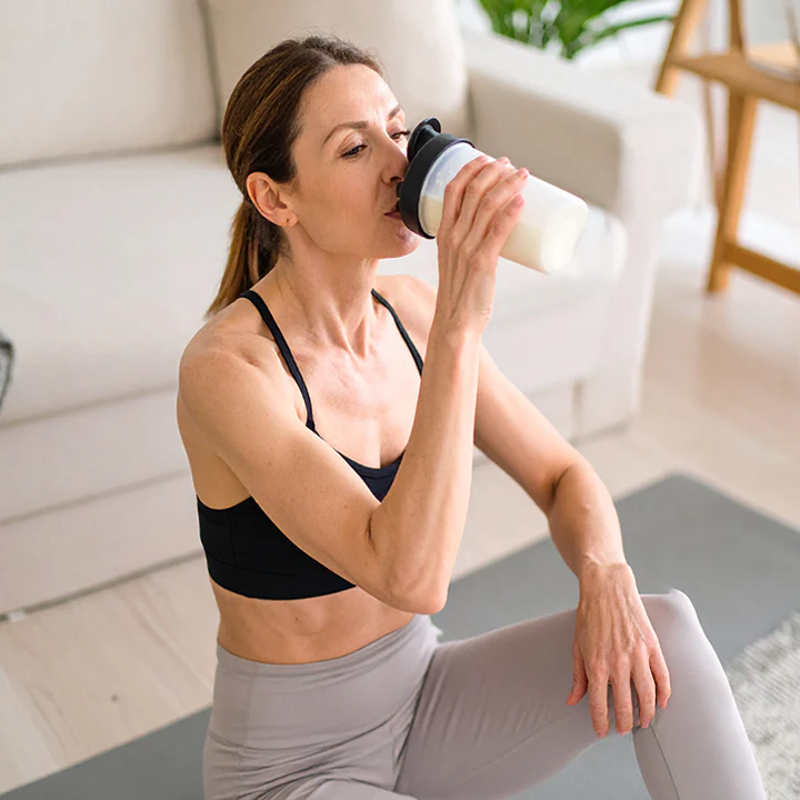 Woman in workout attire drinking from a shaker bottle in a home setting
