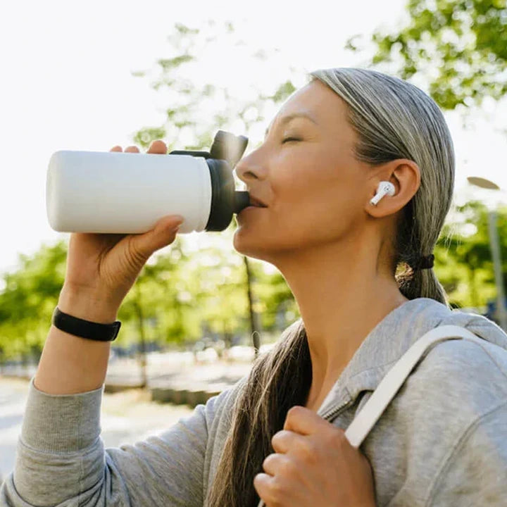 Woman drinking from a water bottle outdoors with trees in the background